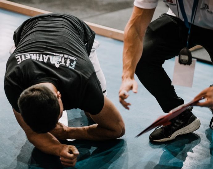 An athlete being assisted by a coach during an indoor training session on a blue floor.