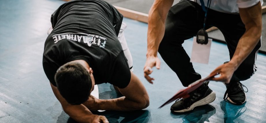 An athlete being assisted by a coach during an indoor training session on a blue floor.